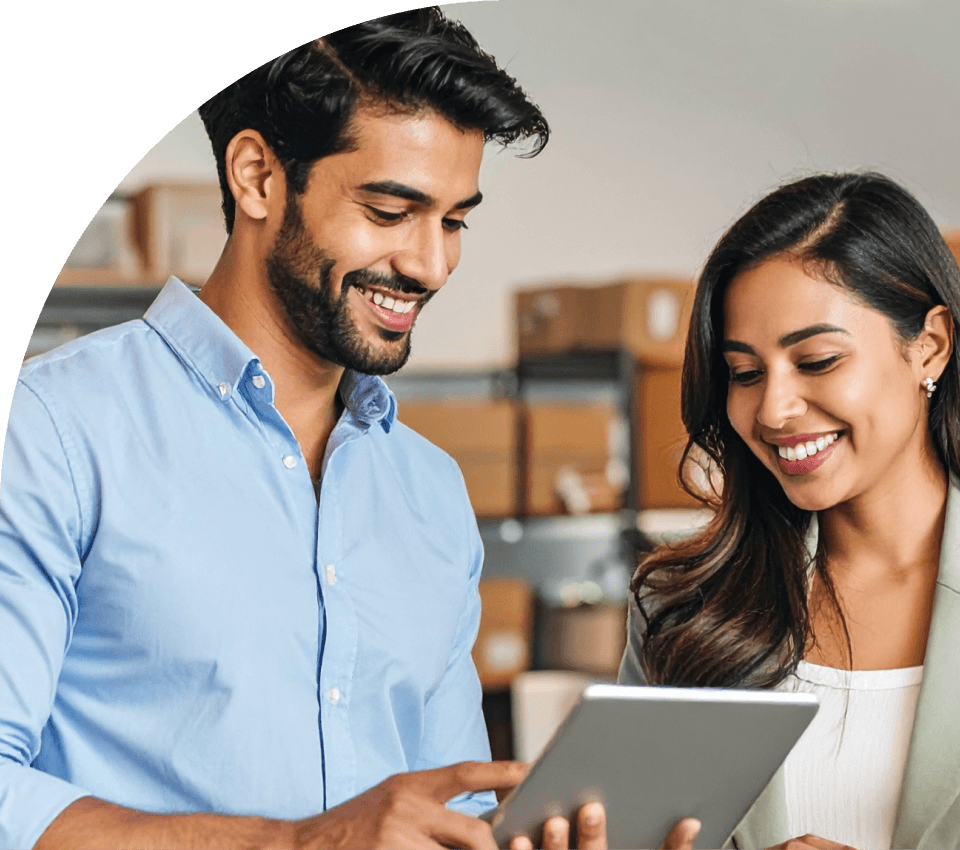 Casal de empreendedores do varejo sorrindo enquanto utilizam um tablet em frente às prateleiras de sua loja, representando a agilidade no tempo de resposta, o aumento das conversões e a transformação do atendimento em um canal de vendas direto.